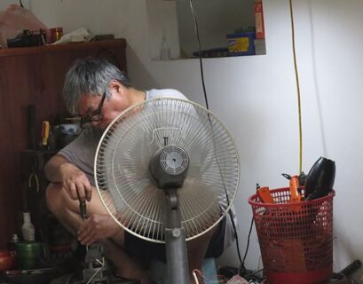 Ein Mann sitzt auf dem Boden und repariert Geräte in einem kleinen Raum mit einem Ventilator.
