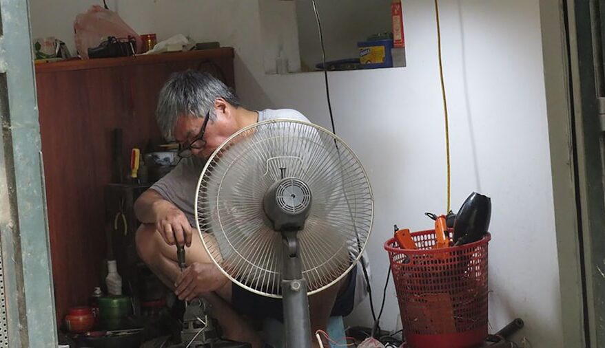 Ein Mann sitzt auf dem Boden und repariert Geräte in einem kleinen Raum mit einem Ventilator.