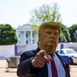 Person trägt eine Donald Trump Maske und zeigt mit dem Finger auf die Kamera vor dem Weißen Haus.