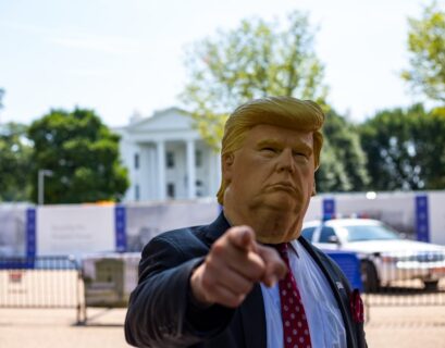 Person trägt eine Donald Trump Maske und zeigt mit dem Finger auf die Kamera vor dem Weißen Haus.