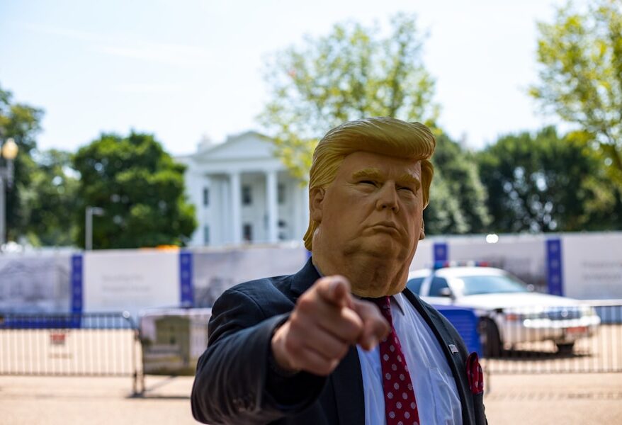 Person trägt eine Donald Trump Maske und zeigt mit dem Finger auf die Kamera vor dem Weißen Haus.