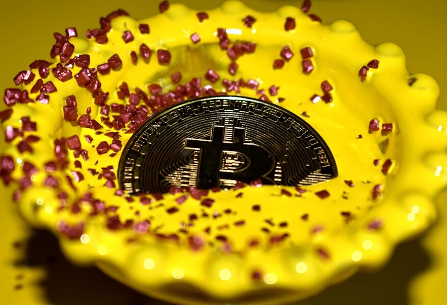 Eine Bitcoin-Münze liegt in einer gelben Schale, umgeben von rosa Zuckerkristallen.