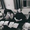 Vier Personen sitzen an einem Tisch während einer Besprechung, mit Unterlagen und Getränken vor ihnen.