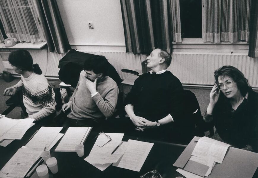 Vier Personen sitzen an einem Tisch während einer Besprechung, mit Unterlagen und Getränken vor ihnen.