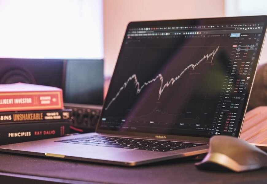MacBook Pro mit einem Finanzdiagramm auf dem Bildschirm und Bücherstapeln im Hintergrund.