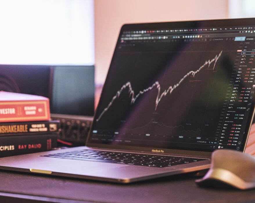 MacBook Pro mit einem Finanzdiagramm auf dem Bildschirm und Bücherstapeln im Hintergrund.